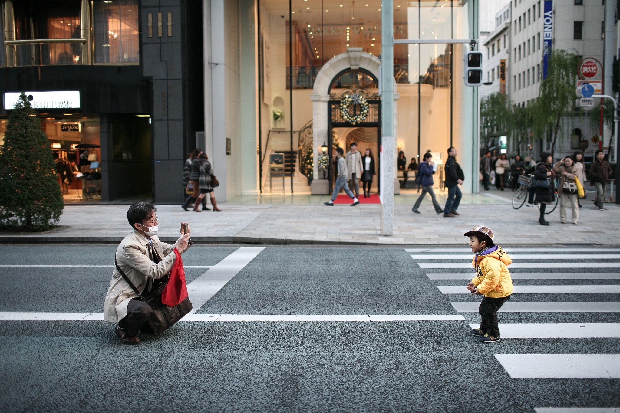 On a Tokyo crosswalk, a man in a trench coat crouches to take a photo of a small child in a yellow jacket.