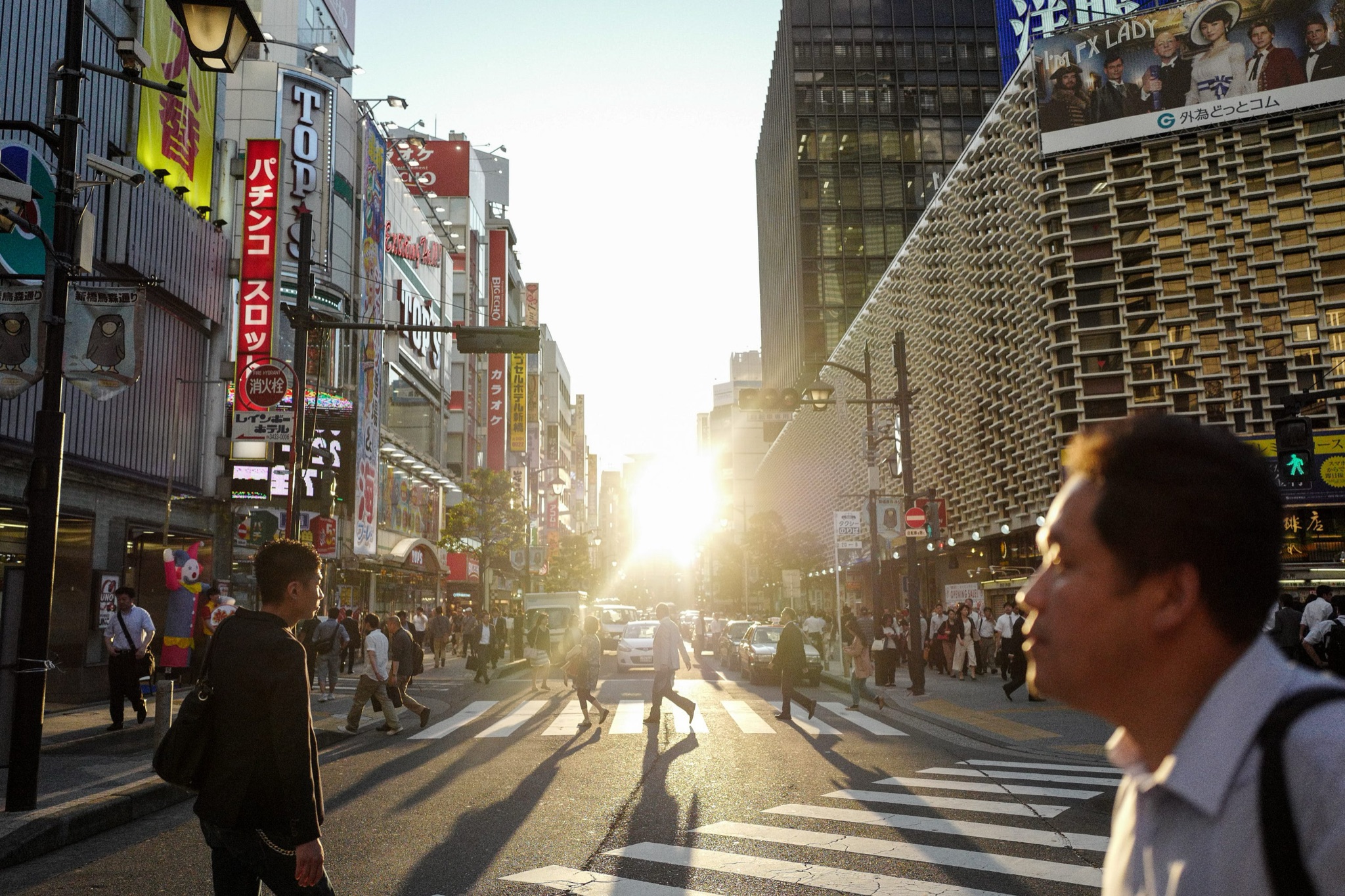 A busy Tokyo street crossing at sunset, with people walking and a man in the foreground.